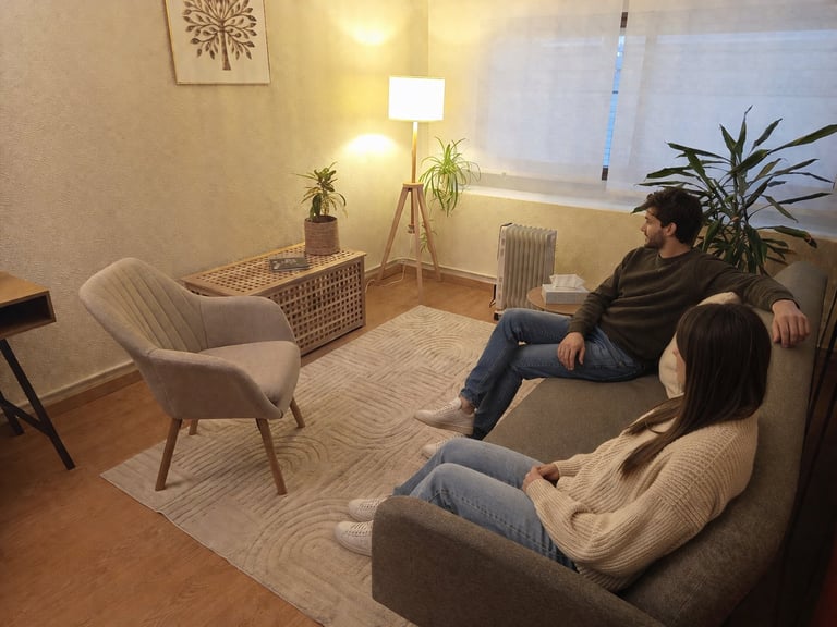 Two people sitting in a cozy living room with armchairs, houseplants, warm lighting, and a window.
